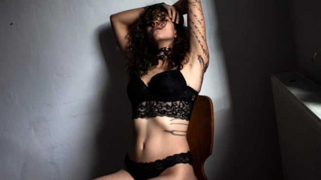 A woman with curly hair poses leaning against a wall in black lace attire, with tattoos visible on her arms and midriff, sitting on a wooden chair.