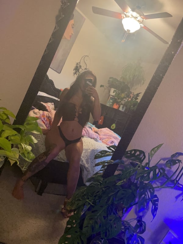 A woman standing in front of a mirror in a bedroom, wearing a swimsuit and smiling while holding her phone.