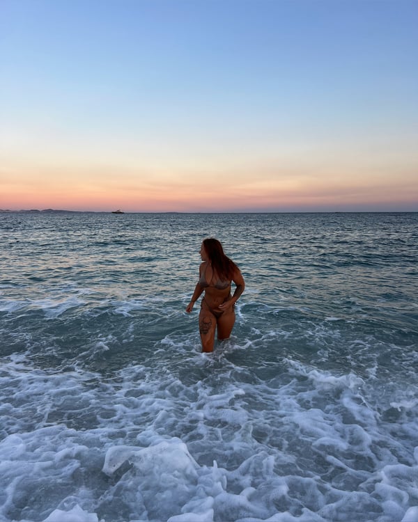 A woman with long red hair stands in shallow ocean water at sunset, wearing a bikini and facing away toward the horizon.