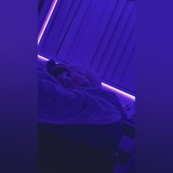 A person lying on a bed with purple neon lighting and blinds in the background.