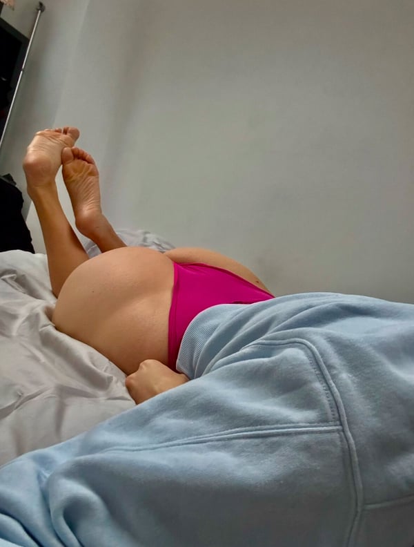 A person lying on a bed in pink underwear, posing with legs raised against a simple indoor background.