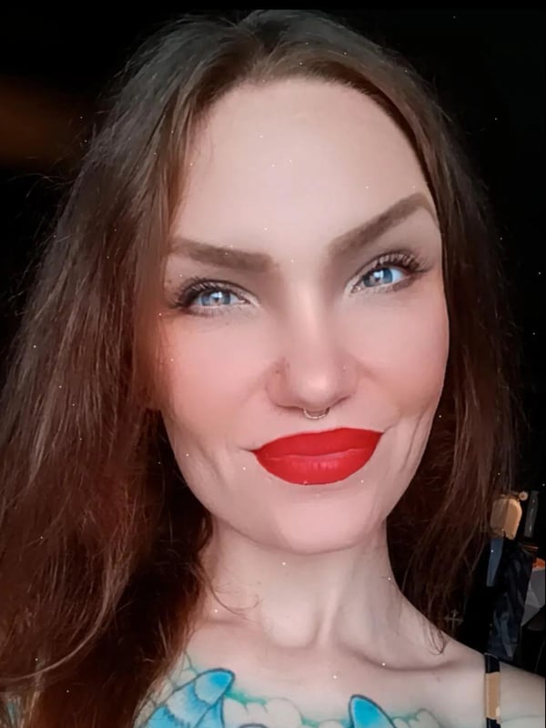 Close-up portrait of a woman with long dark hair, bold makeup, red lips, and visible tattoos on her chest, smiling against a dark background.