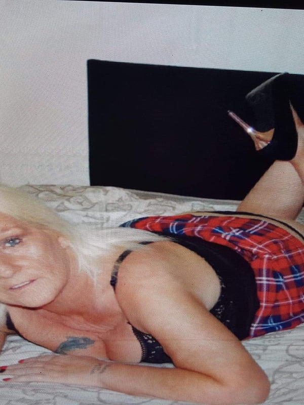 A blonde woman lying on a bed wearing a plaid skirt and black top, with high heels and tattoos visible.