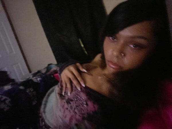 A young woman with long dark hair reclining on a bed with colorful bedding, wearing a patterned top and posing confidently indoors.