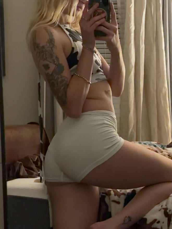 Young woman with long blonde hair taking a mirror selfie in a bikini top, holding a phone, with tattoos on her arm and a beach-themed background.