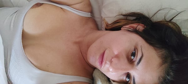 Young woman with long brown hair lying on a bed, wearing a light-colored tank top, gazing sideways at the camera.