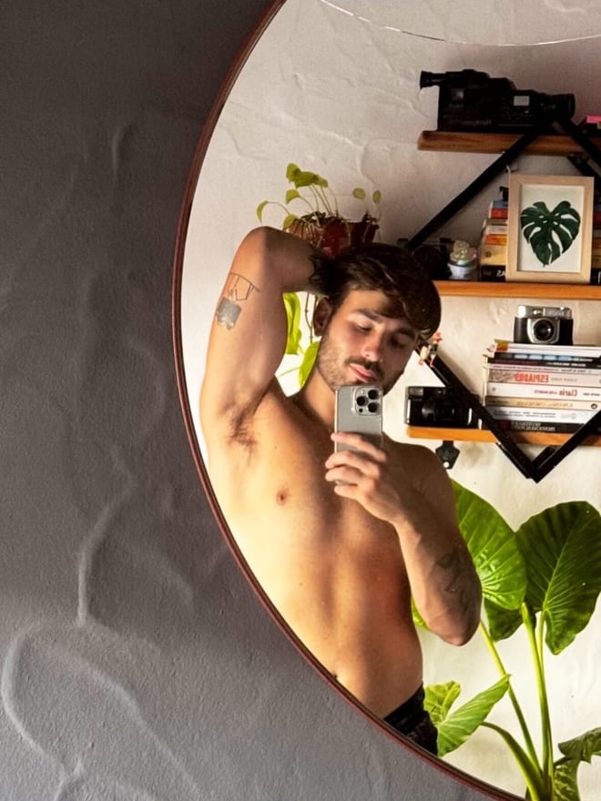 A man taking a mirror selfie in a circular frame, shirtless with tattoos, in a room with plants and decor.