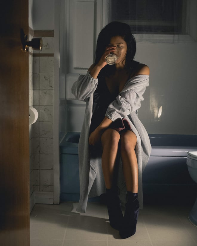 A woman with long dark hair sits on the edge of a bathtub in a dimly lit bathroom, wearing a loose open cardigan over a slip dress and boots, holding a cup near her lips.