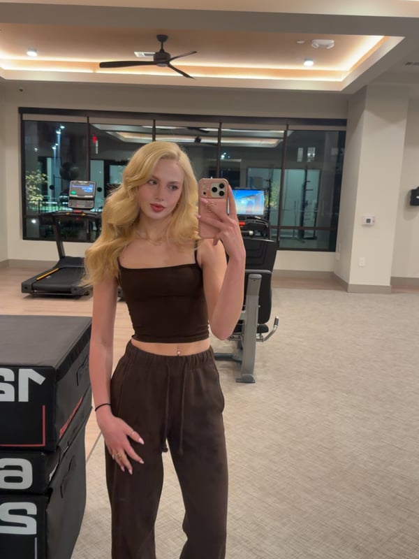 Young woman with blonde hair taking a selfie in a black halter top inside a bedroom with warm lighting.