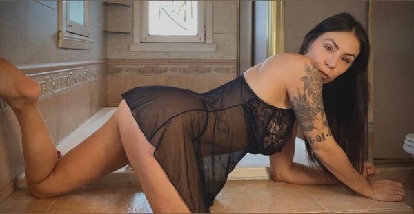 Woman in sheer black lingerie posing seductively on the floor near a bathtub in an indoor setting.