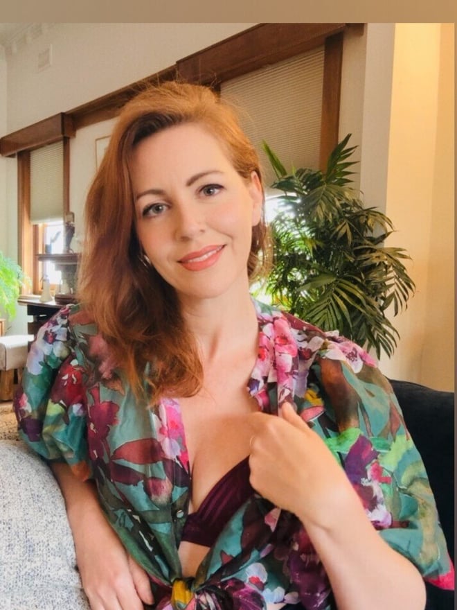 Woman with reddish hair smiling while seated indoors, wearing a colorful floral robe over lingerie, with plants and wooden decor in the background.