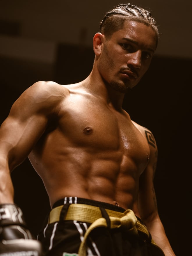 A muscular man with braided hair posing confidently shirtless, wearing martial arts gear and holding gloves.