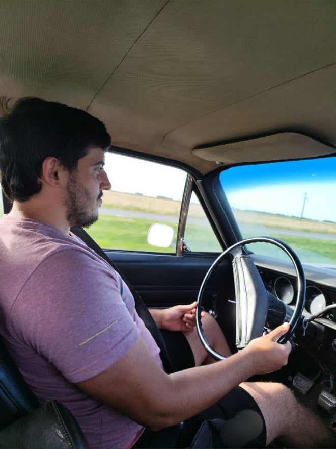 A man with short dark hair sitting in the driver's seat of a vintage car, wearing a pink shirt, with a scenic view outside the window.