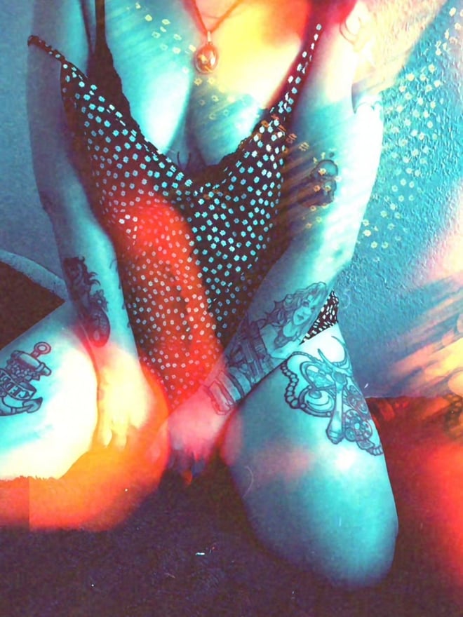 A person sitting in a colorful, glowing outfit with tattoos on arms and legs, necklace visible.