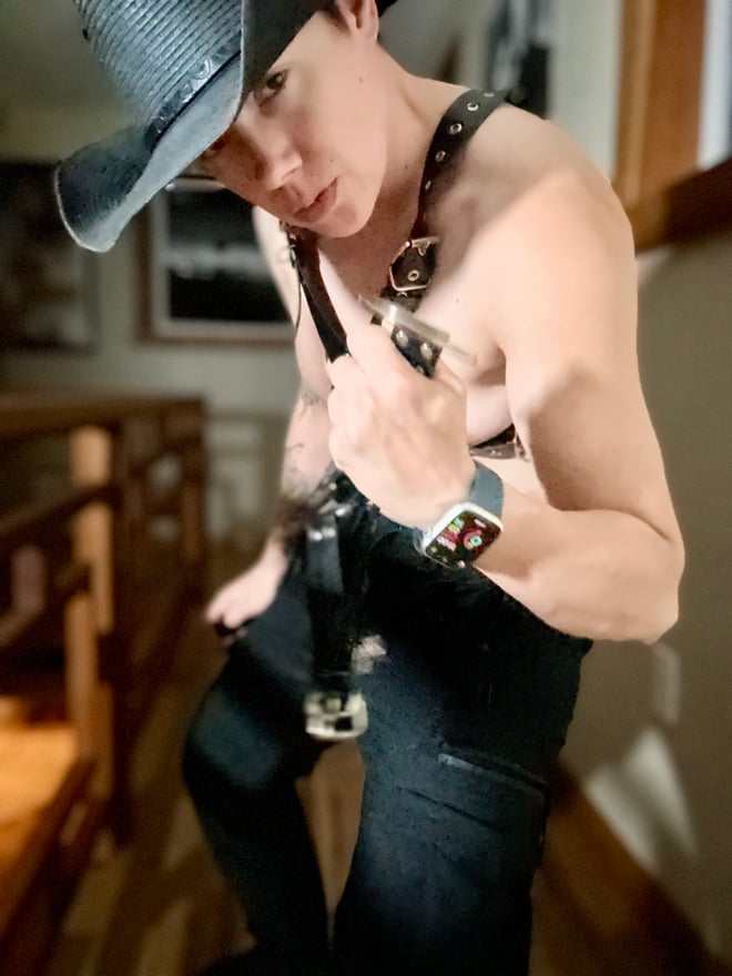 A muscular person in a cowboy hat and dark leather outfit poses against a black background with promotional text.