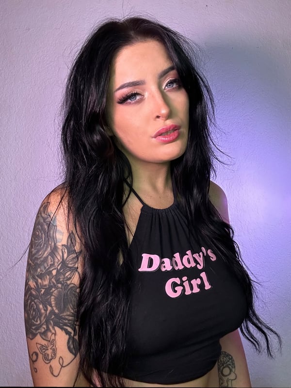 Young woman with long dark hair wearing a black halter top with pink 'Daddy's Girl' text, visible tattoos on arms, posing with a subtle smile against a purple background.