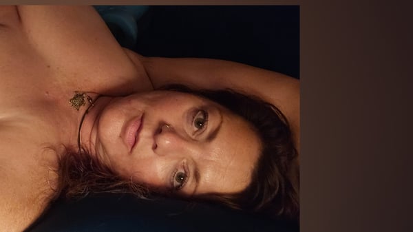 A woman with long hair reclining on a dark surface, wearing a necklace, with a soft expression and warm lighting.
