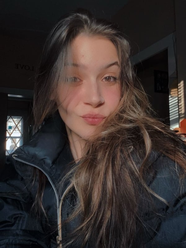 A young woman with long brown hair wearing a black puffer jacket, posing with a slight pout indoors near a window.