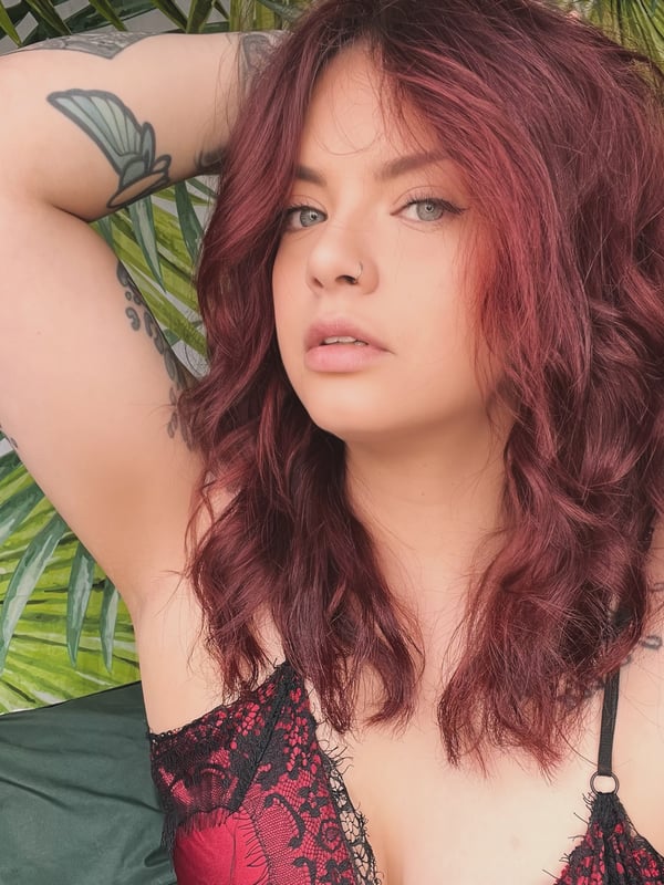 Woman with red wavy hair posing with arm raised against a green leafy background, wearing lace top.