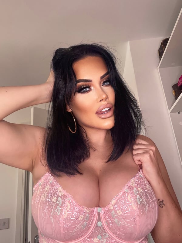 A woman with dark hair and hoop earrings posing in a bedroom setting against a wardrobe, wearing delicate lace attire.