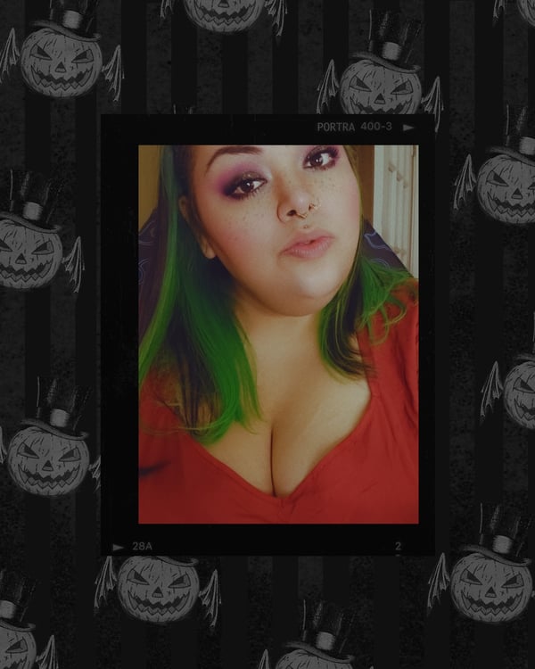 A woman with green hair posing in a red top framed by a black Halloween-themed background with pumpkins.