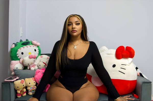 A woman with long highlighted hair sits on a couch surrounded by colorful plush toys, wearing a black bodysuit.