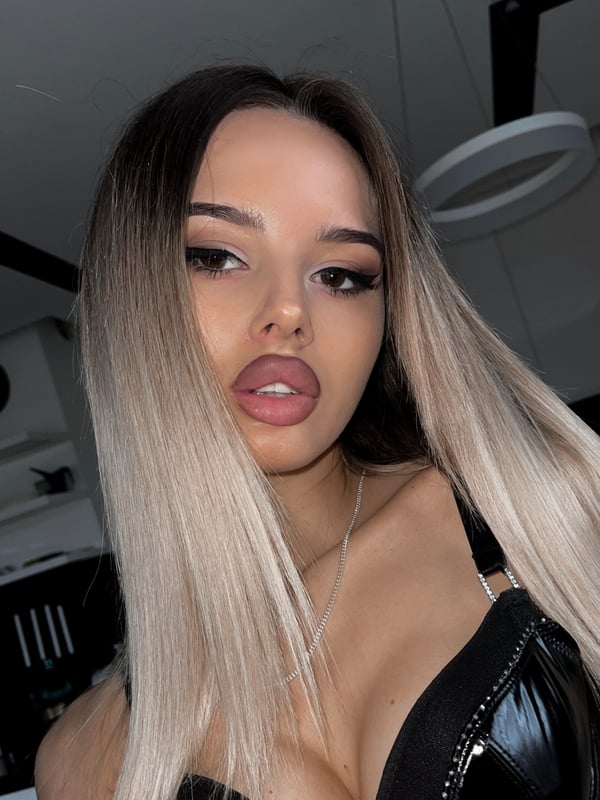 A young woman with long blonde balayage hair, posing confidently indoors with glossy lips and wearing a black top.