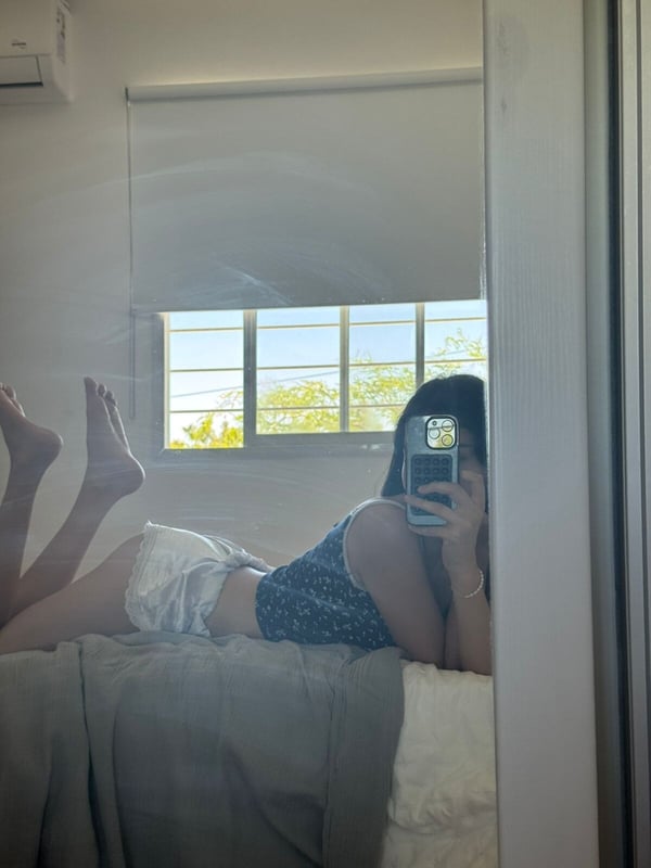 Young woman lying on a bed near a window, taking a mirror selfie while showing her feet.