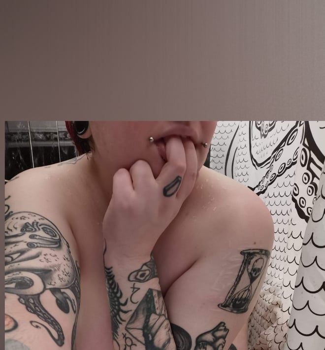 Person with short red hair and facial piercing posing shirtless with hands near mouth against a artistic shower curtain background.