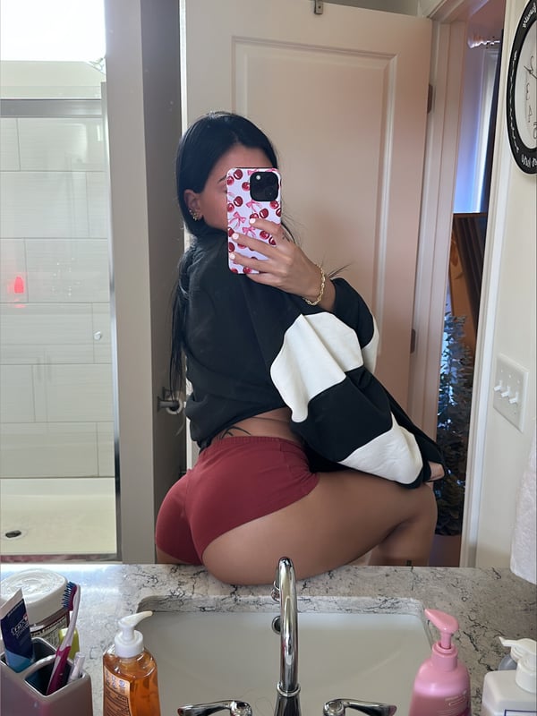 Young woman with dark hair taking a mirror selfie in a bathroom, wearing an oversized black and white shirt with red shorts, smiling playfully.