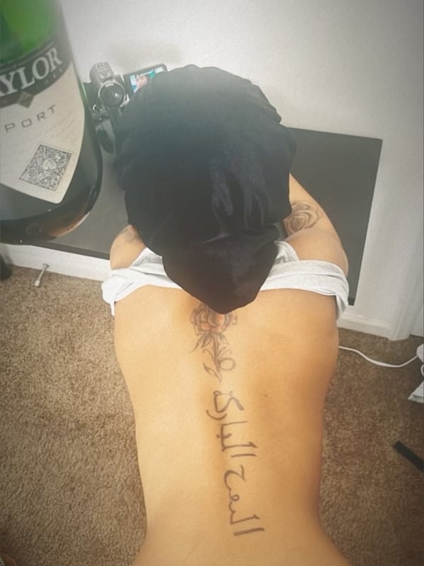 Person with dark hair in a ponytail kneeling on the floor next to a green bottle, showing their back and tattoo in a dimly lit room.