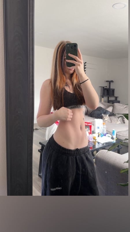 Young woman standing in front of a mirror in a bedroom, wearing a black sports bra and gray sweatpants, posing with her phone.