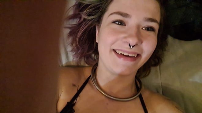 Young woman with colorful hair and piercings smiling at the camera while lying on a bed, wearing a black top and choker necklace.