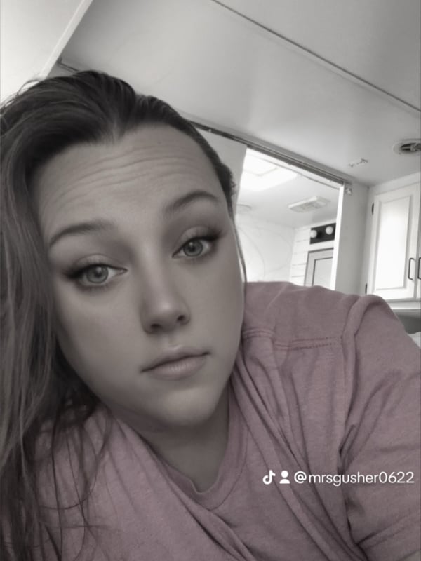 Black and white close-up selfie of a woman with long hair, wearing a pink top, posing indoors near a window.