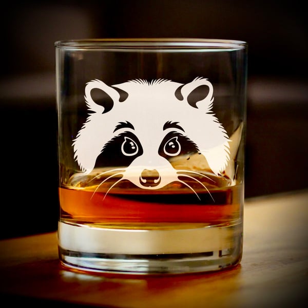 A clear glass tumbler featuring a white silhouette of a raccoon face, containing amber-colored whiskey, placed on a wooden surface.