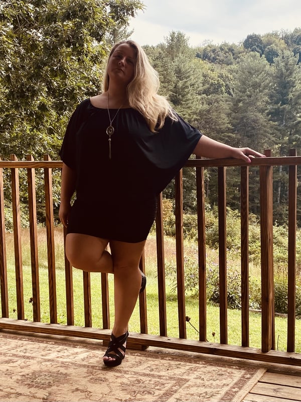 A blonde woman in a black dress standing on a wooden porch railing, smiling outdoors with trees and greenery in the background.