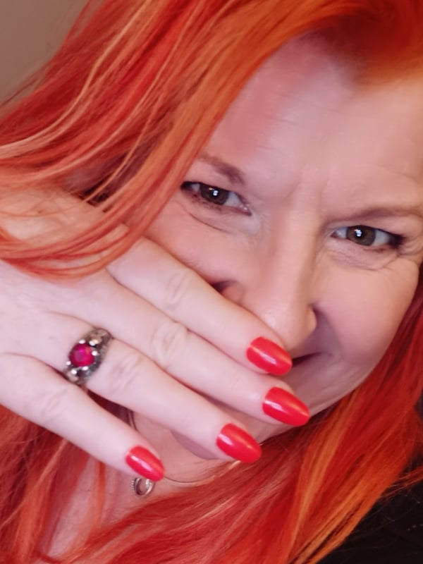 A woman with vibrant red hair smiling playfully while covering part of her face with her hand, featuring long nails.