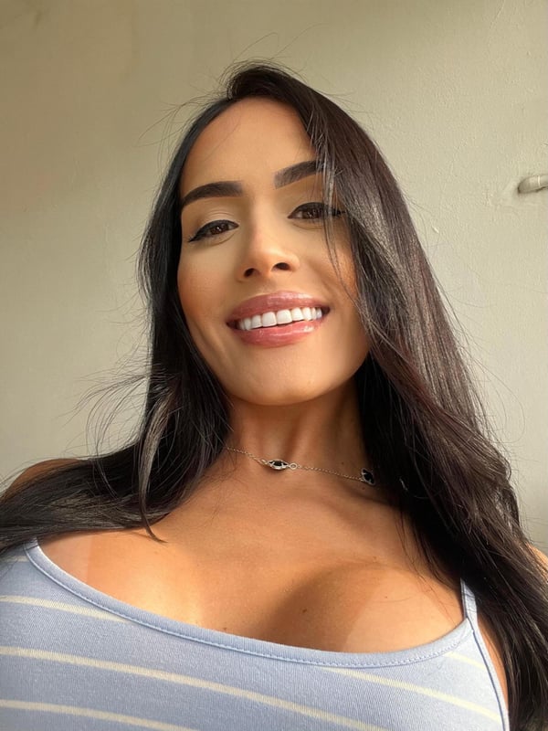 A woman with long dark hair smiling at the camera, wearing a light blue striped top, against a simple indoor background.