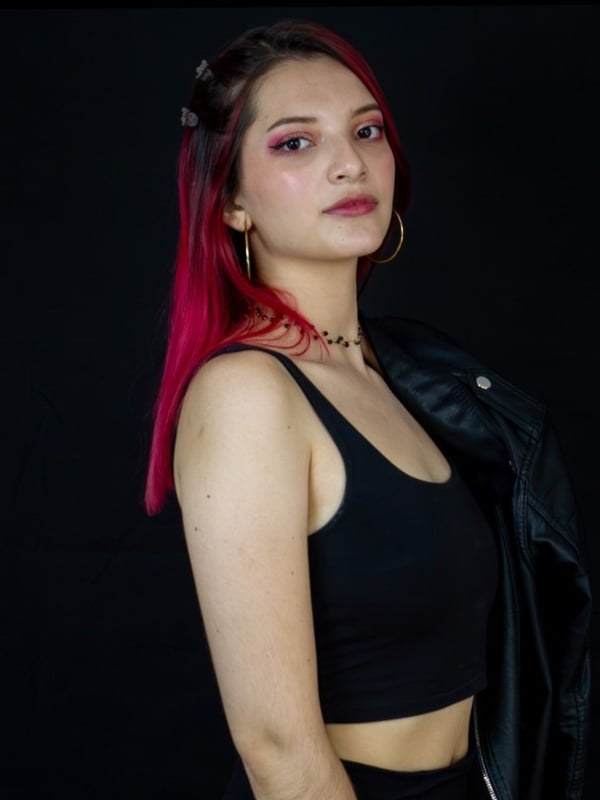 A young woman with pink hair wearing a black tank top and leather jacket, posing against a black background.