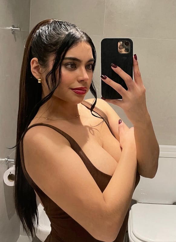 A woman with long dark hair posing on a bed in teal lingerie, resting her head on her hand in a modern bedroom setting.