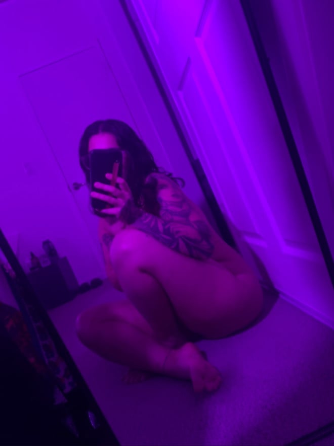 A person with dark hair and tattoos taking a mirror selfie while seated on the floor in a purple-lit room.