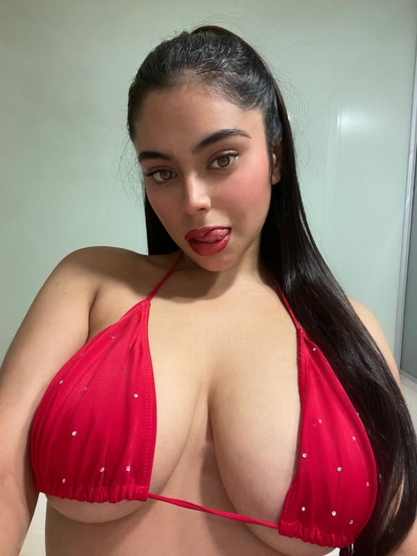 A woman with long dark hair wearing a red lace bra, posing against a plain background while looking at the camera.