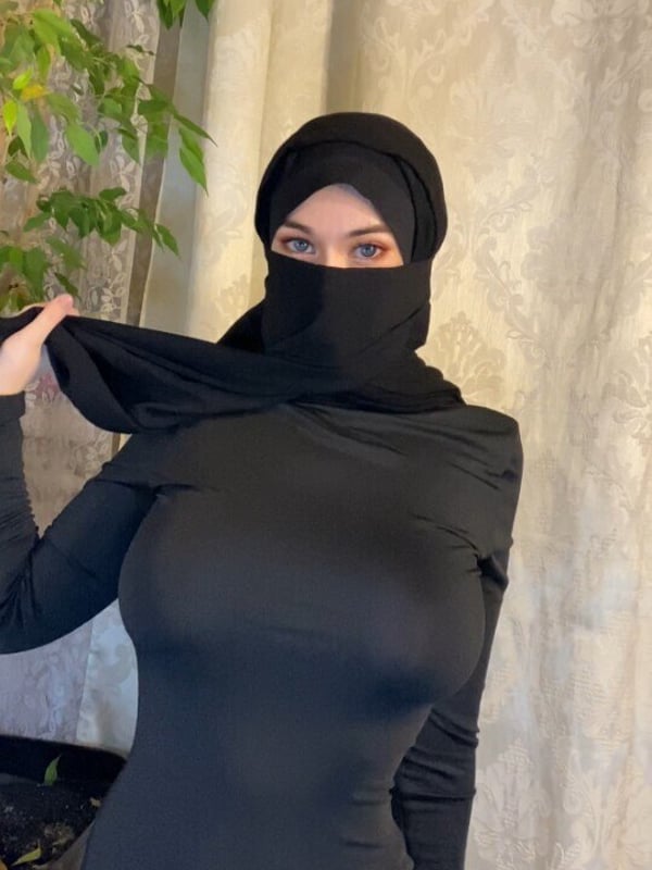 A person wearing a black hijab and face covering, posing indoors near a plant and patterned curtain.