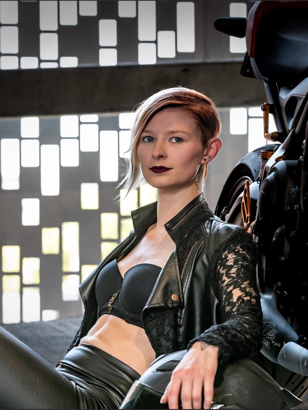 A woman with short blonde hair sitting in a leather jacket and pants next to a motorcycle in a garage setting.