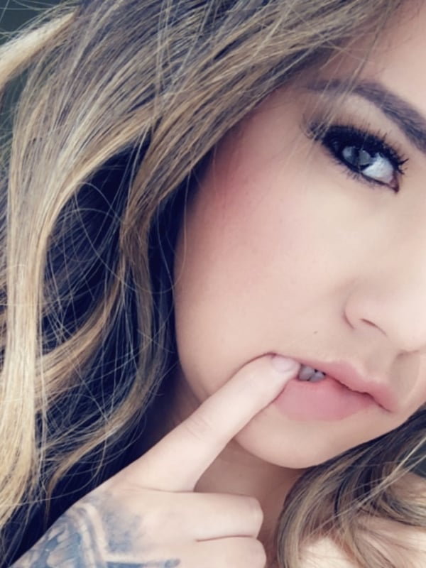 Close-up portrait of a young woman with long wavy hair, touching her lips with her finger, and visible tattoos on her hand.