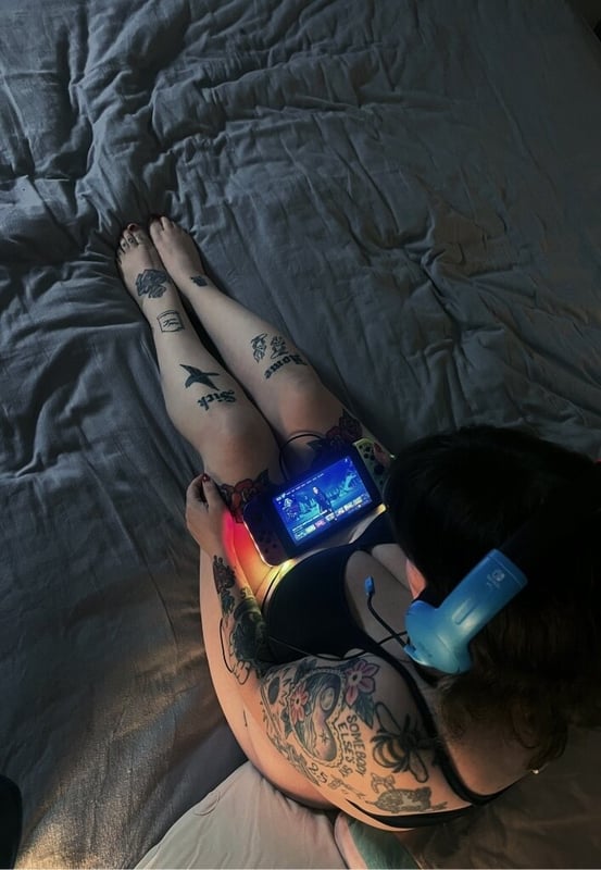 A person lying on a bed playing a handheld gaming console with headphones on, surrounded by tattoos on their legs and arms.