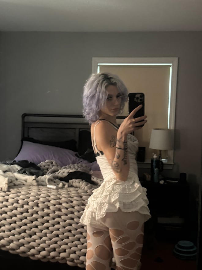 A person with blonde hair, dramatic makeup, facial piercings, and visible tattoos takes a mirror selfie.