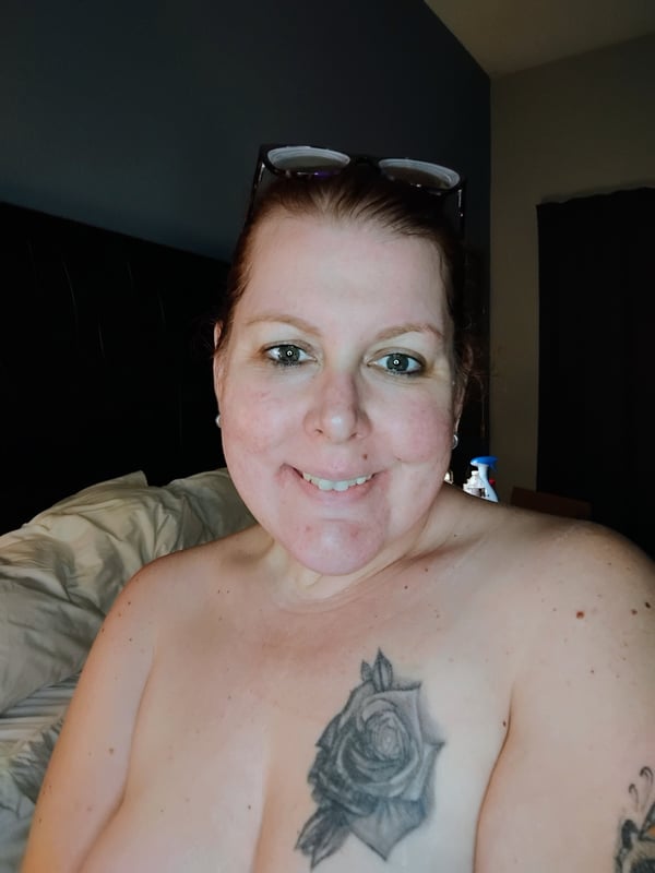 A smiling woman with glasses pushed up on her head and brunette hair, sitting on a bed with a rose tattoo on her chest.