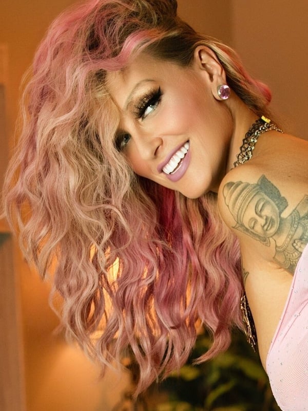 A smiling woman with pink-dyed wavy hair, tattoos, and jewelry poses confidently in a warmly lit indoor setting.
