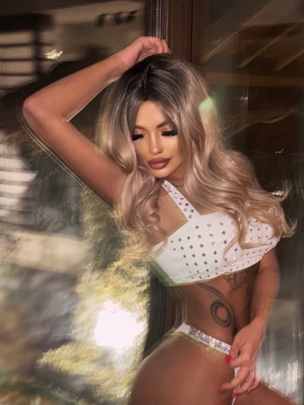 A woman with long blonde hair posing seductively in a white top and bottoms inside a room with a window.